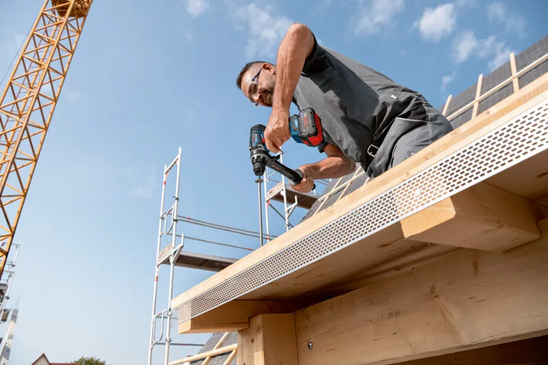 Một người mặc đồ bảo hộ đang khoan vào thanh dầm gỗ của một tòa nhà đang được xây dựng.