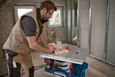 Một người mặc đồ bảo hộ đang cắt gỗ bằng máy cưa bàn không dây tại một khu vực xây dựng.