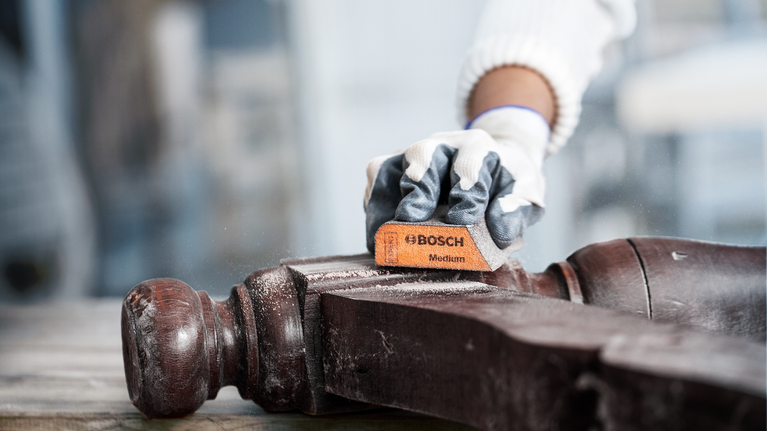 Người mặc đồ bảo hộ đang chà nhám tay vịn ghế bằng gỗ.