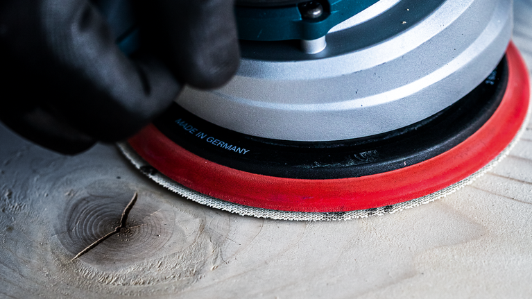 Lưới chà nhám EXPERT M480 cho máy chà nhám quỹ đạo ngẫu nhiên 125 mm, G 120 50 chiếc Người đeo thiết bị an toàn đang chà nhám bề mặt gỗ bằng dụng cụ điện.