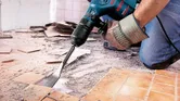 Một người mặc đồ bảo hộ đang dùng búa phá dỡ để tháo gạch lát sàn.