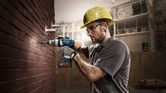 Một người mặc đồ bảo hộ đang khoan vào tường gạch bằng một dụng cụ điện không dây.