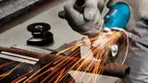 Một người mặc đồ bảo hộ đang cắt một ống kim loại bằng máy mài góc, tia lửa bắn ra.