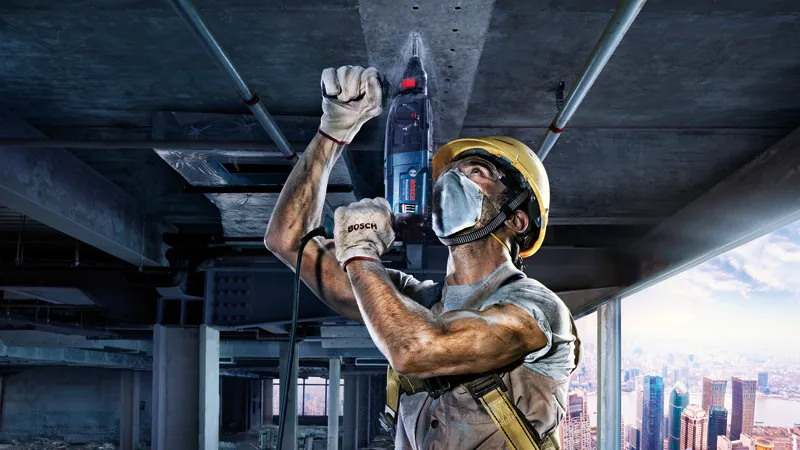 Một người mặc đồ bảo hộ đang khoan trần bê tông tại một công trường xây dựng.