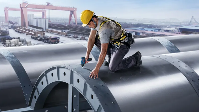 Một người mặc đồ bảo hộ đang dùng dụng cụ điện để siết chặt bu lông trên một kết cấu kim loại lớn.