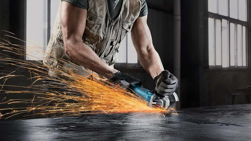 Một người đàn ông mặc đồ bảo hộ đang dùng máy mài góc để mài kim loại, tia lửa bắn ra.