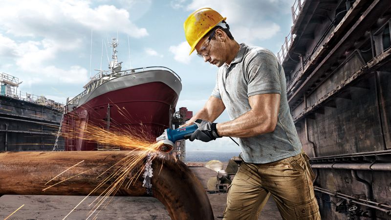 Một người đeo thiết bị an toàn đang mài một ống kim loại, tạo ra tia lửa tại một xưởng đóng tàu.