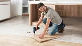 Một người đang khoan vào tấm ván gỗ trên sàn nhà bếp đang được cải tạo.