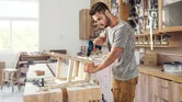 Một người đang lắp ráp một chiếc ghế gỗ bằng máy khoan không dây trong xưởng.