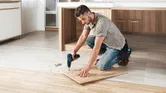 Một người đang khoan vào một tấm ván gỗ trên sàn, xung quanh là vụn gỗ.