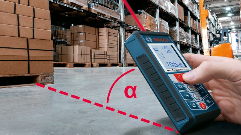 Một người đang sử dụng máy đo laser để kiểm tra khoảng cách trong một nhà kho có các thùng hàng xếp chồng lên nhau.