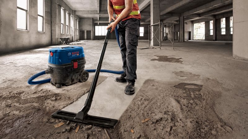 Một người mặc đồ bảo hộ đang hút bụi bê tông trong một tòa nhà lớn chưa hoàn thiện.