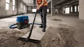 Một người mặc đồ bảo hộ đang hút bụi bê tông trong một tòa nhà lớn chưa hoàn thiện.