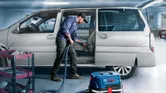 Một người đang vệ sinh nội thất ô tô bằng máy hút bụi bên cạnh xe đẩy đồ nghề trong gara.