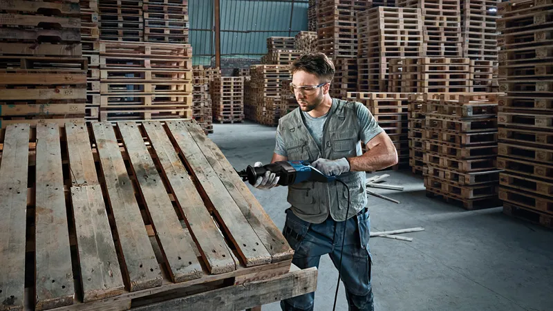 GSA 120 Một người mặc đồ bảo hộ đang cắt một tấm pallet gỗ bằng máy cưa lọng.