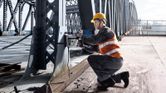 Một người mặc đồ bảo hộ đang khoan vào dầm thép trên cầu.