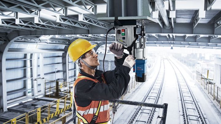 GBM 50-2 Một người mặc đồ bảo hộ đang vận hành máy khoan từ trên một thanh thép phía trên đường ray xe lửa.