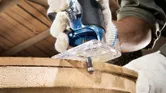 Một người mặc đồ bảo hộ sử dụng máy phay cầm tay để tạo hình cạnh của một tấm ván gỗ.