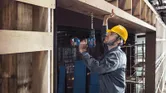Một người mặc đồ bảo hộ đang khoan vào thanh dầm gỗ tại một công trường xây dựng.