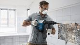 Một người mặc đồ bảo hộ đang sử dụng búa phá dỡ để phá vỡ gạch ốp tường.
