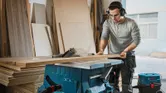 Một người mặc đồ bảo hộ đang cắt tấm gỗ bằng máy cưa bàn trong xưởng.
