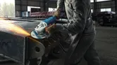Một người mặc đồ bảo hộ đang mài một thanh kim loại lớn bằng máy mài góc.