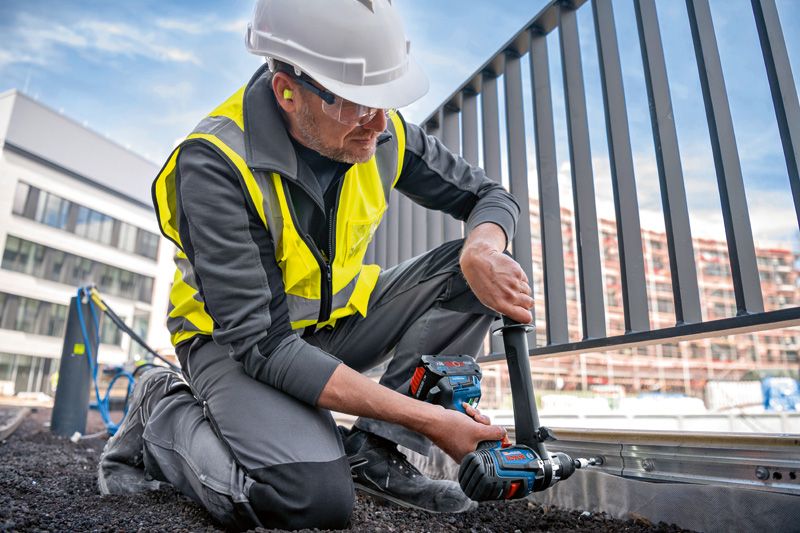 Một người mặc đồ bảo hộ đang sử dụng máy khoan không dây trên khung hàng rào kim loại.