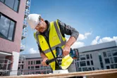 Một người mặc đồ bảo hộ đang khoan lỗ vào gỗ tại một công trường xây dựng.