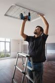 Một người mặc đồ bảo hộ đang lắp đèn trần bằng máy khoan điện.