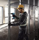 Một người mặc đồ bảo hộ sẽ cố định các đinh kim loại bằng máy khoan không dây.