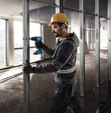 Một người mặc đồ bảo hộ sẽ cố định các đinh kim loại bằng máy khoan không dây.