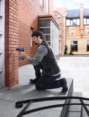 Một người mặc đồ bảo hộ đang khoan vào bức tường gạch ở bên ngoài tòa nhà.