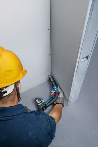 Một người mặc đồ bảo hộ đang cố định vách thạch cao bằng tua vít không dây.