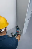 Một người mặc đồ bảo hộ đang cố định vách thạch cao bằng tua vít không dây.