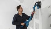 Một người đang chà nhám tường thạch cao bằng máy chà nhám tường thạch cao trên bức tường chưa hoàn thiện.