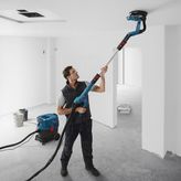 Một người mặc đồ bảo hộ đang chà nhám trần nhà bằng máy chà nhám tường thạch cao được kết nối với máy hút bụi.