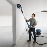 Một người đang chà nhám bức tường trắng bằng máy chà nhám tường thạch cao được kết nối với máy hút bụi.