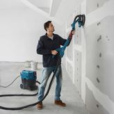 Một người đang chà nhám tường thạch cao bằng máy chà nhám tường thạch cao được kết nối với máy hút bụi.