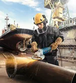 Một người mặc đồ bảo hộ đang mài một ống kim loại lớn tại xưởng đóng tàu.