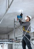 Một người mặc đồ bảo hộ đang khoan trần bê tông bằng búa quay không dây.