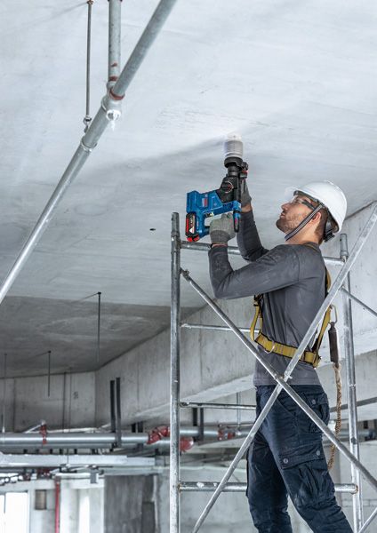 Một người mặc thiết bị an toàn đứng trên giàn giáo, sử dụng máy khoan điện trên trần nhà.