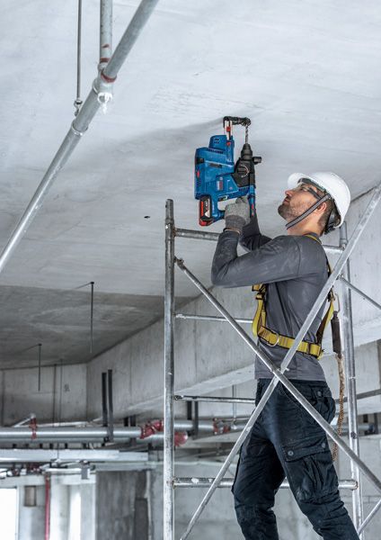 Một người mặc đồ bảo hộ đang khoan vào trần bê tông trong khi đứng trên giàn giáo.