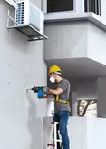 Một người mặc đồ bảo hộ đang khoan lỗ trên tường bê tông trong khi đứng trên thang.
