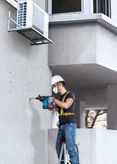 Một người mặc đồ bảo hộ đang khoan vào bức tường bê tông trong khi đứng trên thang.
