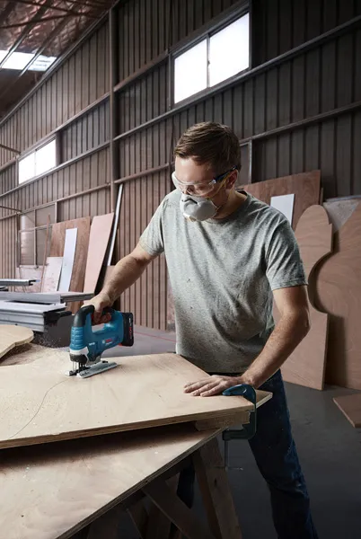 Một người mặc đồ bảo hộ đang cắt ván ép bằng máy cưa lọng không dây trong xưởng.