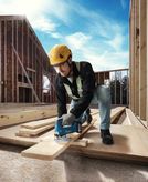 Một người mặc đồ bảo hộ đang cắt ván gỗ bằng máy cưa lọng không dây tại một công trường xây dựng.