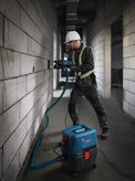 Một người mặc đồ bảo hộ đang khoan tường, sử dụng máy hút bụi ướt/khô để hút bụi.