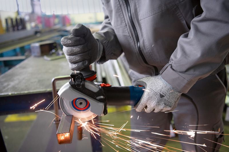 Một người đeo thiết bị an toàn đang mài một thanh kim loại bằng máy mài góc cầm tay.