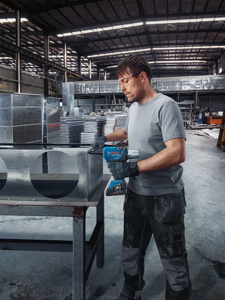 Một người mặc thiết bị an toàn sẽ cố định ống gió kim loại bằng súng bắn đinh tán không dây.