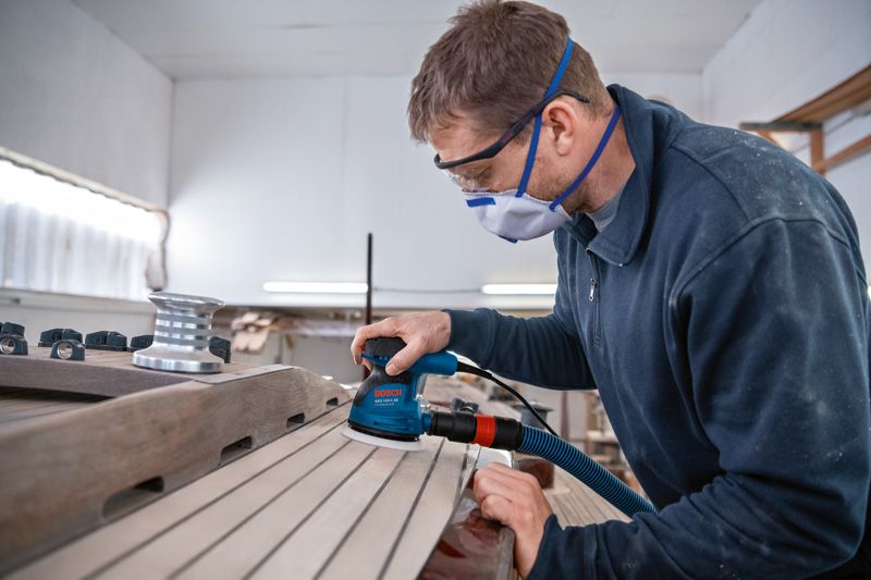 Một người mặc đồ bảo hộ đang chà nhám ván gỗ bằng máy chà nhám quỹ đạo ngẫu nhiên.